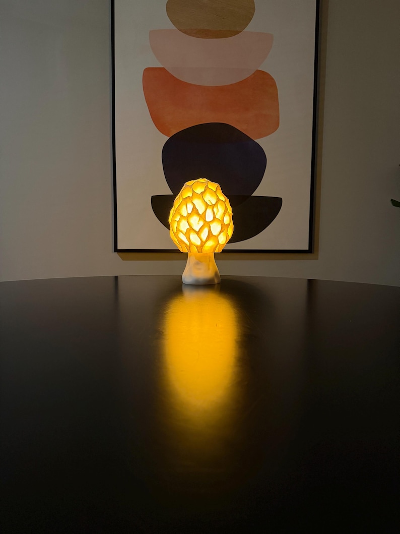 Puede incluir: Una l&aacute;mpara con forma de seta con una tapa texturizada similar a un panal que emite un c&aacute;lido resplandor amarillo dorado. La base es lisa y blanca, y la luz proyecta un reflejo brillante y alargado sobre la superficie oscura debajo.