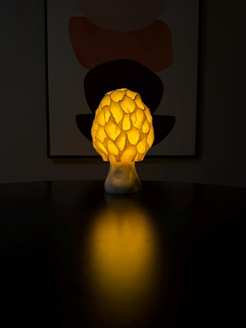 Puede incluir: Una l&aacute;mpara de mesa con forma de seta que presenta una c&uacute;pula texturizada tipo panal que brilla con una luz amarilla c&aacute;lida. La base es lisa y blanca, sosteniendo la parte superior iluminada que proyecta un suave resplandor dorado.