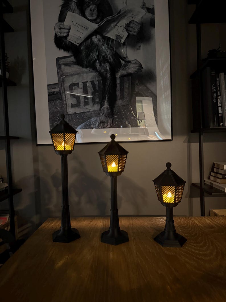May include: Three black street lamp-style lanterns of varying heights, each featuring a hexagonal base, a vertical post, and a lantern head with a diamond-patterned mesh enclosure. Each lantern contains a warm, glowing yellow light inside.