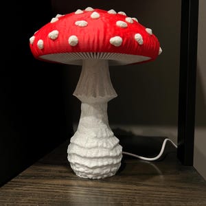 May include: A mushroom-shaped lamp featuring a vibrant red cap with raised white spots and a textured white stalk. The underside of the cap displays visible gills, and a thin white power cord extends from the base of the lamp to provide electricity.