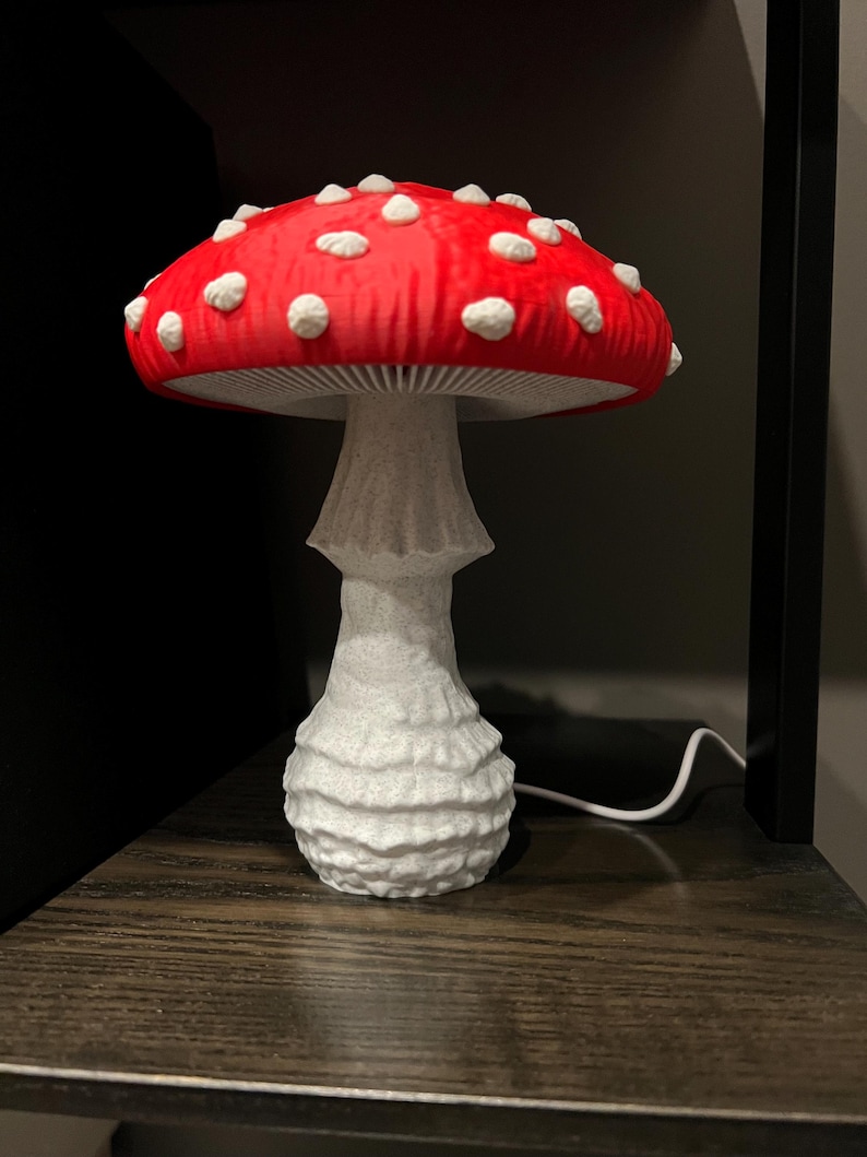 May include: A mushroom-shaped lamp featuring a vibrant red cap with raised white spots and a textured white stalk. The underside of the cap displays visible gills, and a thin white power cord extends from the base of the lamp to provide electricity.