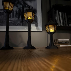 May include: Three black, miniature street lamps of varying heights, each featuring a hexagonal lantern head with a lattice pattern and a warm, glowing yellow light inside. The lamps have fluted posts and square bases, standing upright.