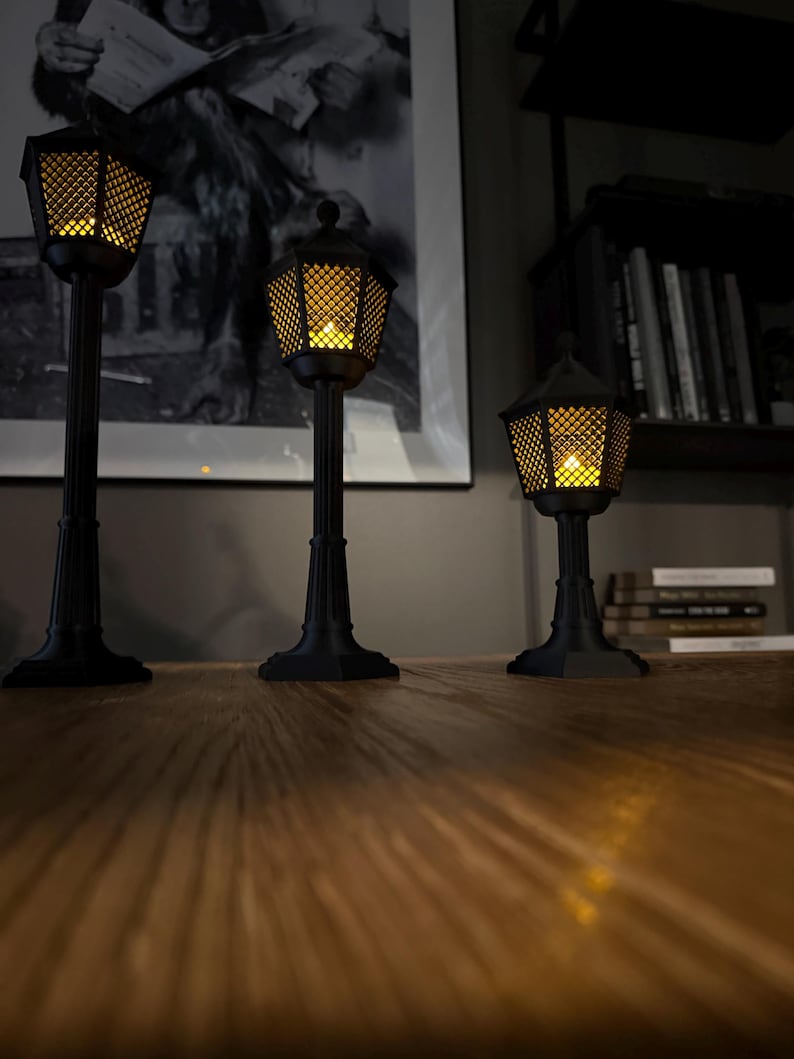 May include: Three black, miniature street lamps of varying heights, each featuring a hexagonal lantern head with a lattice pattern and a warm, glowing yellow light inside. The lamps have fluted posts and square bases, standing upright.