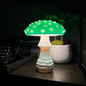 May include: A mushroom-shaped lamp featuring a vibrant green cap with raised, lighter-colored spots. The stem is textured with a white, ridged upper section that transitions into a brown, earthy base. The lamp emits a soft, warm glow from underneath.