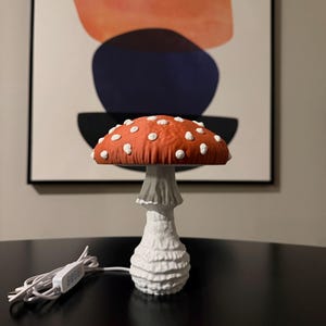 May include: A mushroom-shaped lamp featuring a textured orange cap with raised white spots and a white, ridged stalk. The lamp includes a white power cord with an inline switch, showcasing a detailed, organic design that mimics a toadstool.