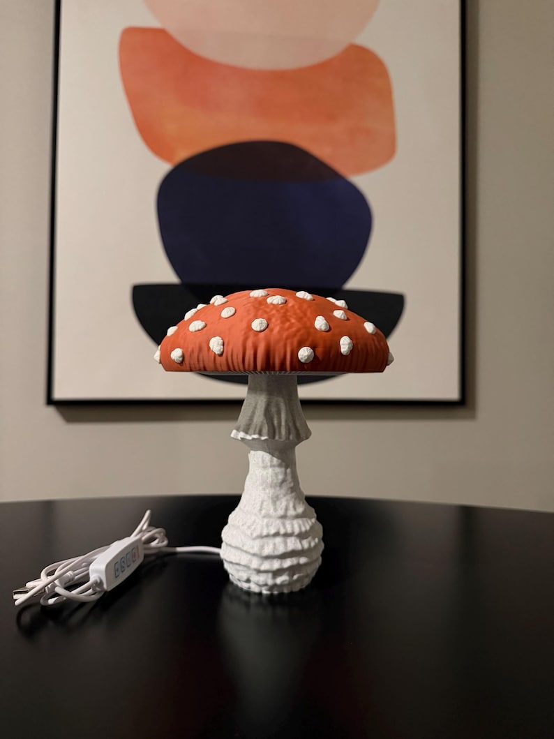 May include: A mushroom-shaped lamp featuring a textured orange cap with raised white spots and a white, ridged stalk. The lamp includes a white power cord with an inline switch, showcasing a detailed, organic design that mimics a toadstool.