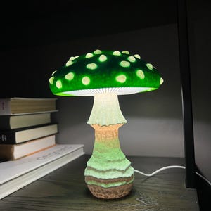 May include: A mushroom-shaped table lamp featuring a vibrant green cap with raised white spots. The underside of the cap is illuminated with white light, casting a glow down onto the textured, light-colored stem that transitions to a brown base.