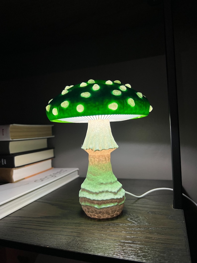 May include: A mushroom-shaped table lamp featuring a vibrant green cap with raised white spots. The underside of the cap is illuminated with white light, casting a glow down onto the textured, light-colored stem that transitions to a brown base.