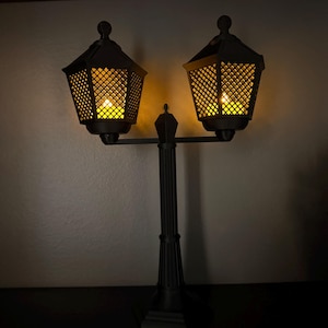 May include: A black double-headed lamp post with a central vertical column and two hexagonal lanterns. Each lantern features a lattice-patterned frame and emits a warm, glowing yellow light from within, illuminating the surrounding area.