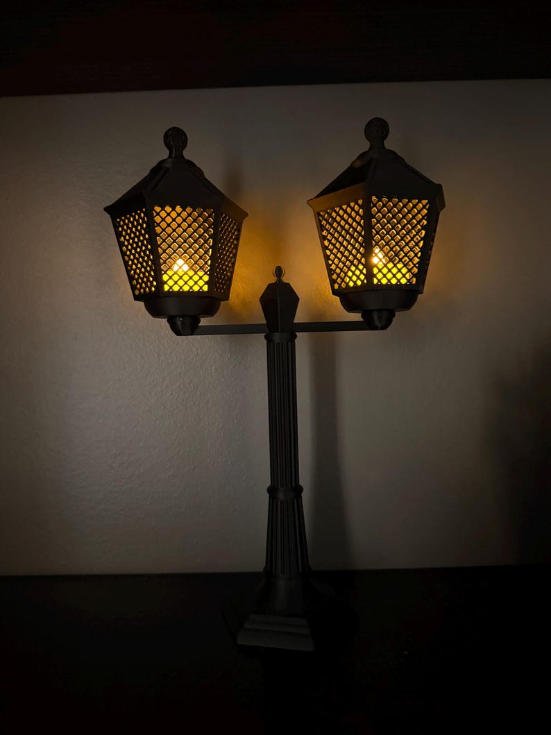 May include: A black double-headed lamp post with a central vertical column and two hexagonal lanterns. Each lantern features a lattice-patterned frame and emits a warm, glowing yellow light from within, illuminating the surrounding area.