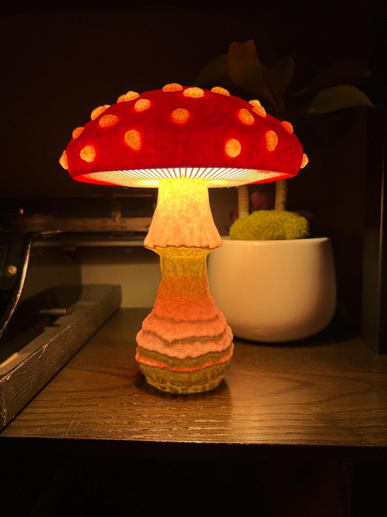 May include: A mushroom-shaped lamp with a vibrant red cap featuring raised, glowing yellow spots. The stem is textured with horizontal ridges and emits a warm, golden light that illuminates the underside of the cap and the surrounding area.