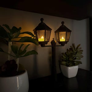 May include: A dark-coloured double lamp post decoration featuring two hexagonal lanterns with a lattice pattern and glowing yellow lights, positioned between two small potted green plants in white ceramic containers.
