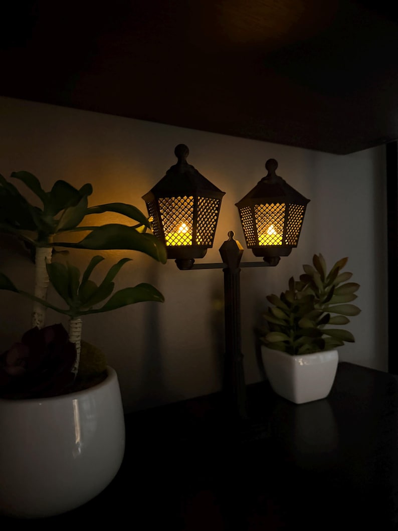 May include: A dark-coloured double lamp post decoration featuring two hexagonal lanterns with a lattice pattern and glowing yellow lights, positioned between two small potted green plants in white ceramic containers.