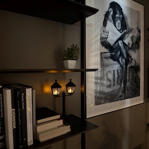 May include: A black metal bookshelf featuring a small decorative lamp with two lantern-style shades, a potted succulent, and a collection of books. Beside the shelf hangs a large framed black and white print of a chimpanzee reading a newspaper.