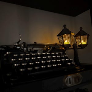 May include: A black vintage typewriter with circular keys and a metallic carriage sits next to a decorative dual-lamp post fixture. The lamps feature a dark lattice frame and emit a warm, glowing yellow light, illuminating the typewriter keys.