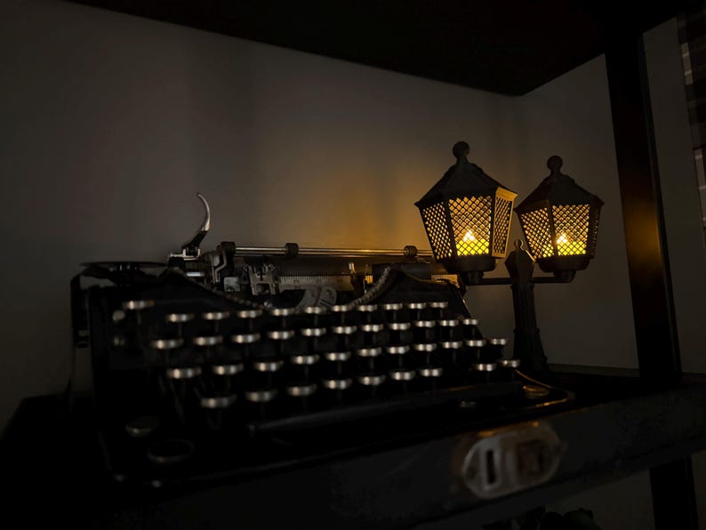 May include: A black vintage typewriter with circular keys and a metallic carriage sits next to a decorative dual-lamp post fixture. The lamps feature a dark lattice frame and emit a warm, glowing yellow light, illuminating the typewriter keys.