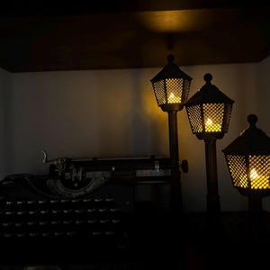 May include: A vintage black typewriter sits beside three dark, post-style lamps of varying heights. Each lamp features a hexagonal, lattice-patterned shade with a warm, glowing light inside, creating a soft illumination in the dark setting.