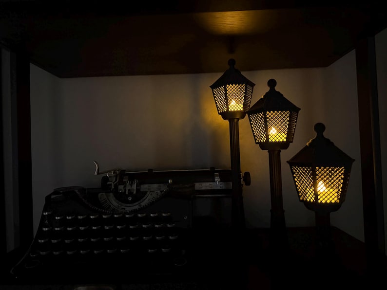 May include: A vintage black typewriter sits beside three dark, post-style lamps of varying heights. Each lamp features a hexagonal, lattice-patterned shade with a warm, glowing light inside, creating a soft illumination in the dark setting.