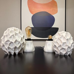 Puede incluir: Dos l&aacute;mparas de mesa blancas con pantallas de textura org&aacute;nica similar a un panal y bases redondeadas y lisas. Cada l&aacute;mpara cuenta con una bombilla expuesta de estilo Edison con filamento visible, que emite un brillo c&aacute;lido sobre el blanco.