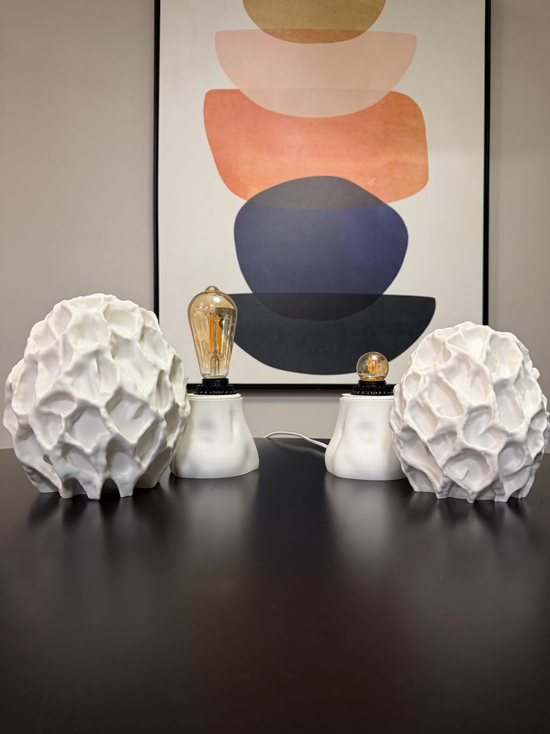 Puede incluir: Dos l&aacute;mparas de mesa blancas con pantallas de textura org&aacute;nica similar a un panal y bases redondeadas y lisas. Cada l&aacute;mpara cuenta con una bombilla expuesta de estilo Edison con filamento visible, que emite un brillo c&aacute;lido sobre el blanco.