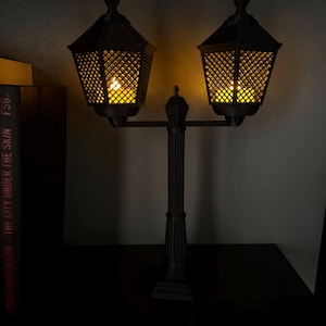 May include: A black double-headed lamp post with a central column and two hexagonal lanterns featuring a lattice pattern. Each lantern emits a warm yellow glow from within, illuminating the dark structure against a plain background.