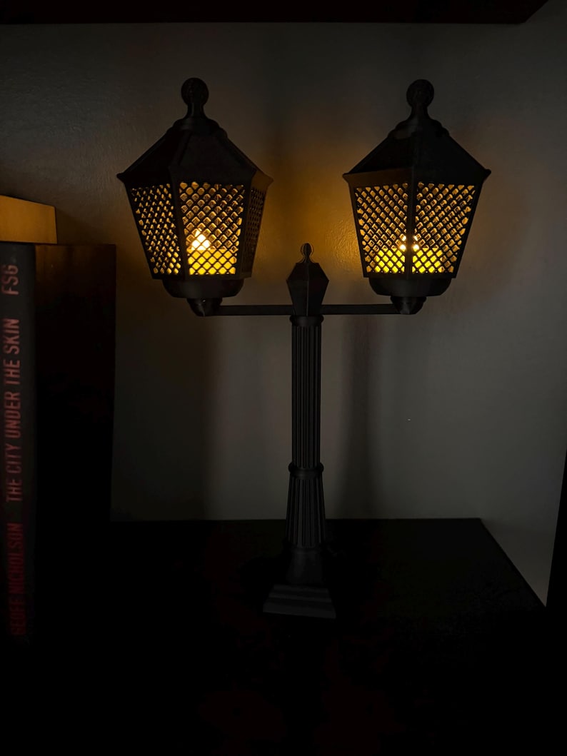 May include: A black double-headed lamp post with a central column and two hexagonal lanterns featuring a lattice pattern. Each lantern emits a warm yellow glow from within, illuminating the dark structure against a plain background.