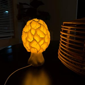 Puede incluir: Una l&aacute;mpara de mesa con forma de seta que presenta una c&uacute;pula texturizada similar a un panal, la cual emite una c&aacute;lida luz de color amarillo dorado. La l&aacute;mpara cuenta con una base s&oacute;lida y lisa y un cable de alimentaci&oacute;n blanco.