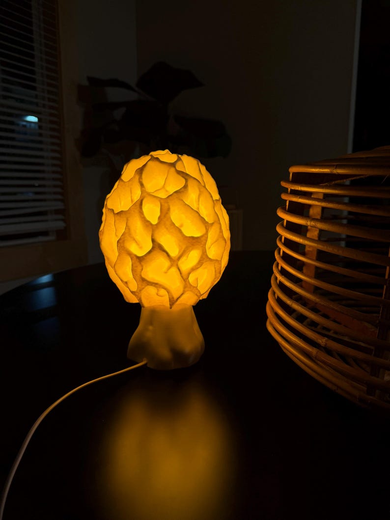 Puede incluir: Una l&aacute;mpara de mesa con forma de seta que presenta una c&uacute;pula texturizada similar a un panal, la cual emite una c&aacute;lida luz de color amarillo dorado. La l&aacute;mpara cuenta con una base s&oacute;lida y lisa y un cable de alimentaci&oacute;n blanco.