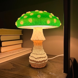 May include: A mushroom-shaped table lamp featuring a vibrant green cap with raised, light-colored circular spots. The stem has a textured, off-white to tan gradient finish. The lamp emits a warm, soft glow from beneath the cap, illuminating the stem.