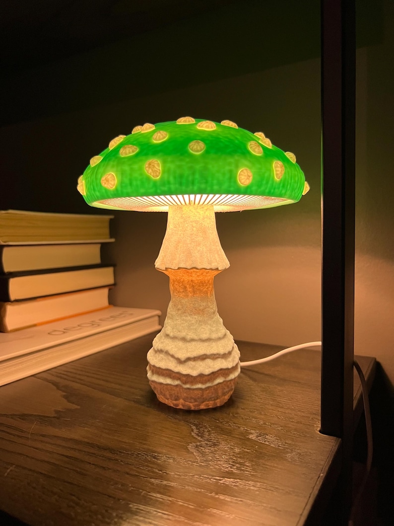 May include: A mushroom-shaped table lamp featuring a vibrant green cap with raised, light-colored circular spots. The stem has a textured, off-white to tan gradient finish. The lamp emits a warm, soft glow from beneath the cap, illuminating the stem.