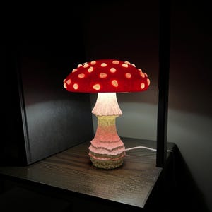 May include: A mushroom-shaped table lamp featuring a vibrant red cap with raised, glowing white spots. The stem has a textured, layered appearance with a gradient of soft pink and pale yellow tones, emitting a warm light from beneath the cap.