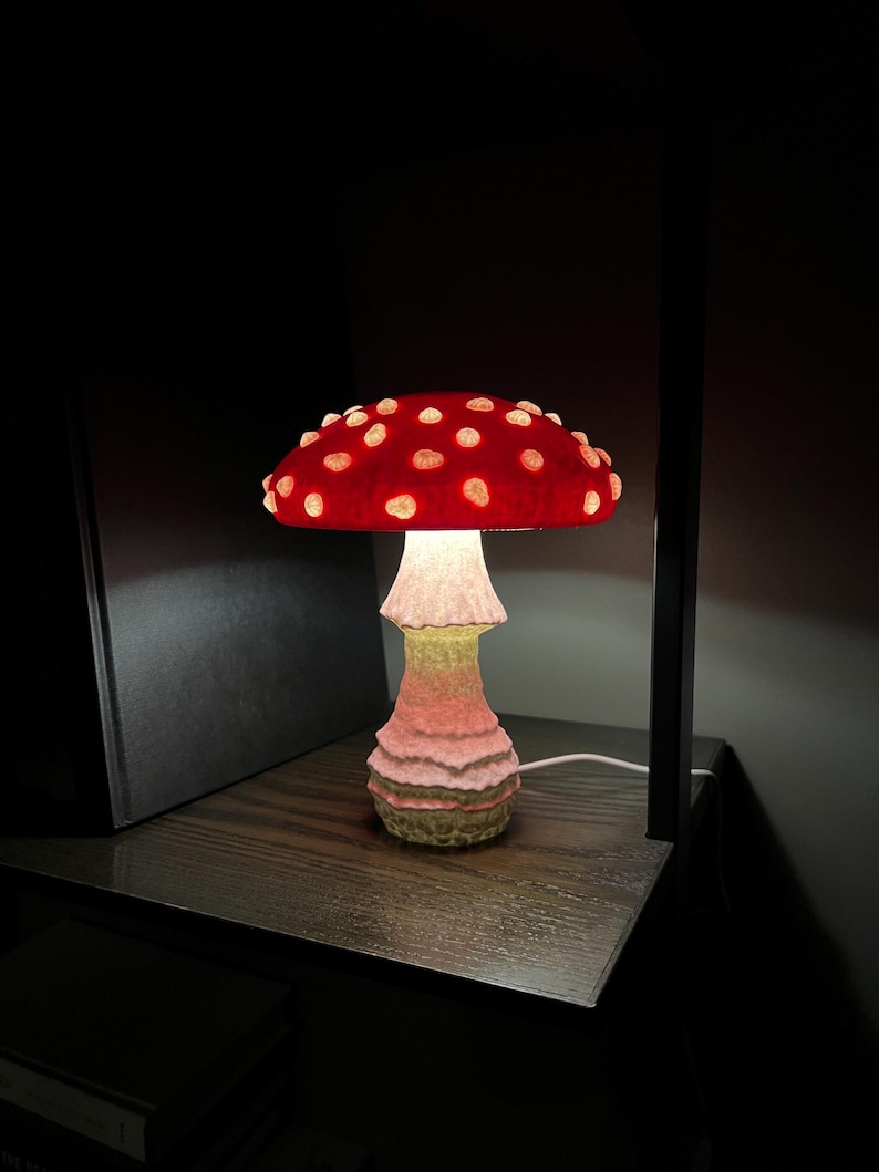 May include: A mushroom-shaped table lamp featuring a vibrant red cap with raised, glowing white spots. The stem has a textured, layered appearance with a gradient of soft pink and pale yellow tones, emitting a warm light from beneath the cap.