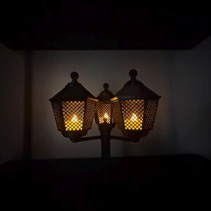 May include: A dark-coloured, three-headed lantern fixture featuring a lattice-patterned metal frame. Each of the three hexagonal lamp heads contains a warm, glowing light source, casting a soft illumination from within the intricate, crisscross metal design.