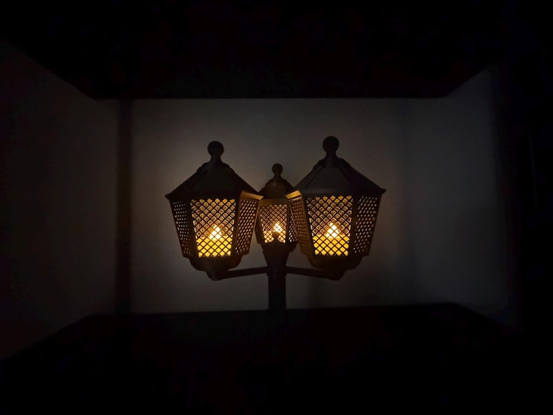 May include: A dark-coloured, three-headed lantern fixture featuring a lattice-patterned metal frame. Each of the three hexagonal lamp heads contains a warm, glowing light source, casting a soft illumination from within the intricate, crisscross metal design.