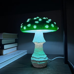 May include: A mushroom-shaped lamp emitting a vibrant green glow from its cap and a softer white light from its stem. The cap features raised, lighter-colored spots, while the stem has a textured, uneven surface. A white power cord extends from the base.