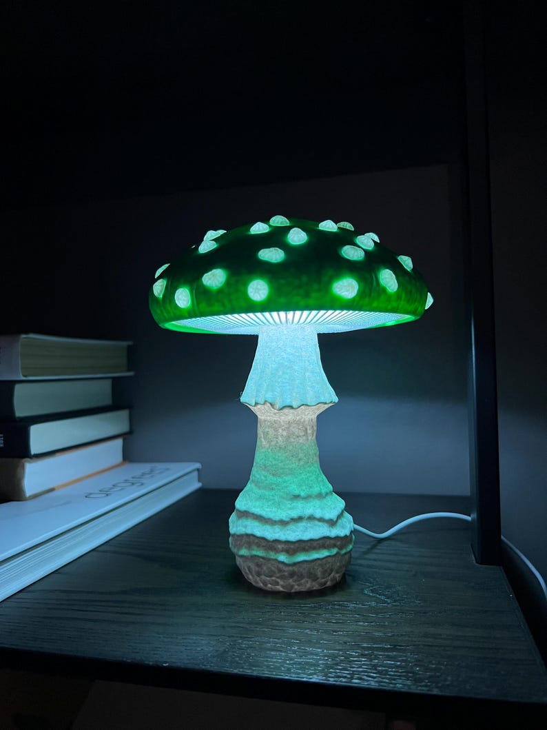 May include: A mushroom-shaped lamp emitting a vibrant green glow from its cap and a softer white light from its stem. The cap features raised, lighter-colored spots, while the stem has a textured, uneven surface. A white power cord extends from the base.