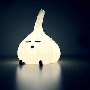 May include: A whimsical, illuminated garlic bulb-shaped lamp. The white lamp has a textured surface, black eyes, a dot for a mouth, and two small black feet. The lamp is lit from within, casting a soft glow.