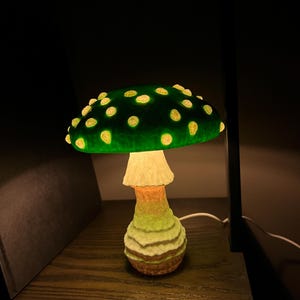 May include: A mushroom-shaped lamp featuring a dark green cap with glowing yellow spots. The stem has a textured, layered appearance with gradient colors transitioning from light cream at the top to a brownish hue at the base, emitting a soft light.