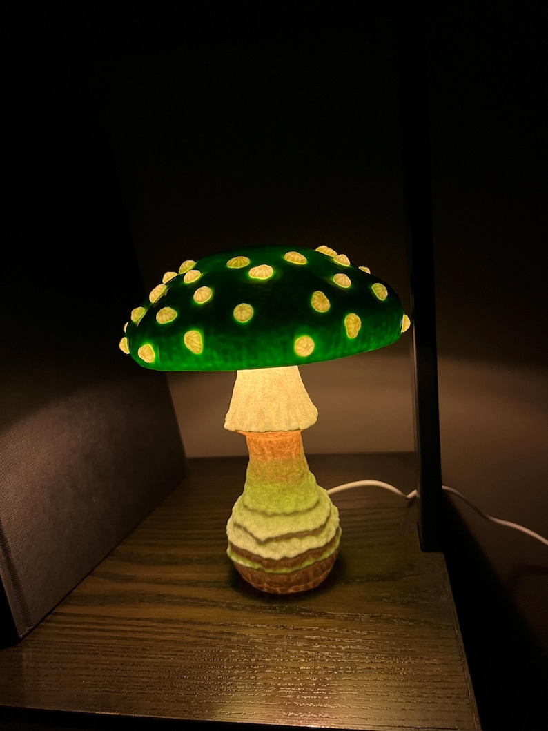 May include: A mushroom-shaped lamp featuring a dark green cap with glowing yellow spots. The stem has a textured, layered appearance with gradient colors transitioning from light cream at the top to a brownish hue at the base, emitting a soft light.