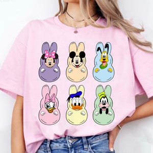 Peut inclure: T-shirt rose clair avec six personnages de dessins animés déguisés en lapins de Pâques. Les personnages incluent Minnie Mouse, Mickey Mouse, Pluto, Daisy Duck, Donald Duck et Dingo. Le design est coloré et ludique.