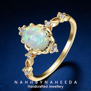 May include: A gold ring featuring an oval opal gemstone with iridescent colors. The ring is accented with small, clear gemstones and decorative gold detailing. The text "NAHHBYNAHEEDA Handcrafted Jewellery" is at the bottom.