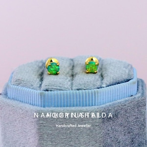 May include: A pair of gold stud earrings with vibrant green opal stones. The earrings are displayed on a blue velvet jewelry box. The text "NAMQBRYNUASHTALDA Handcrafted Jeweller" is visible at the bottom of the image.