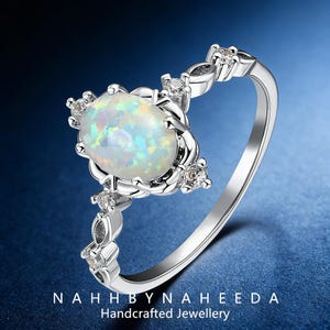 May include: A silver ring featuring an oval opal gemstone as the centerpiece, surrounded by smaller clear gemstones. The ring's band has a delicate design with additional small gemstones. The text "NAHHBYNAHEEDA Handcrafted Jewellery" is at the bottom.