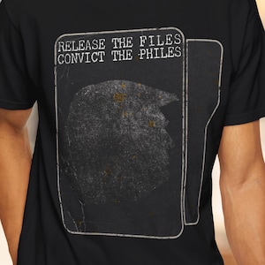 Puede incluir: Una camiseta negra de manga corta con un gráfico blanco que muestra el contorno de una carpeta de archivos que contiene la silueta oscura de una cabeza. El texto "RELEASE THE FILES CONVICT THE PHILES" está impreso en letras blancas.