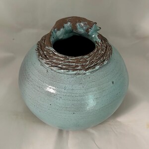 May include: A handcrafted ceramic vase with a round body and a textured, woven-like rim. The vase is a light blue-green color with a brown accent around the opening. The unique design adds a touch of artistry to any space.