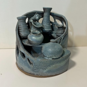 May include: A blue-grey ceramic sculpture featuring a tiered structure with multiple small vases and pots integrated into a curved, open-work frame. The piece has a textured, speckled glaze finish and includes a small sculpted figure of a cat.
