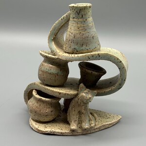 May include: A unique ceramic sculpture featuring a cat figurine and several small, textured vases. The sculpture has a speckled, earthy color palette with shades of brown, beige, and a hint of blue-green. The design includes curved elements and multiple levels.
