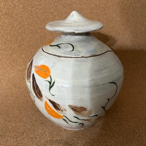 May include: A white ceramic jar with a lid. The jar is decorated with orange and brown floral designs and green stems. The lid is cone-shaped. The jar is approximately 8 inches tall.