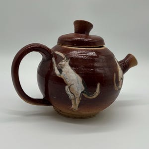 May include: A handmade, brown ceramic teapot with a lid and handle. The teapot features a painted white cat design on the side. The spout and handle are a darker shade of brown.