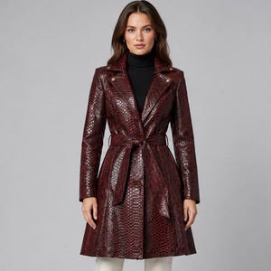 May include: A dark burgundy trench coat featuring a textured snakeskin pattern, a wide notched collar, long sleeves, and a matching tie belt at the waist. The coat is worn over a black turtleneck sweater and is paired with light-colored trousers.